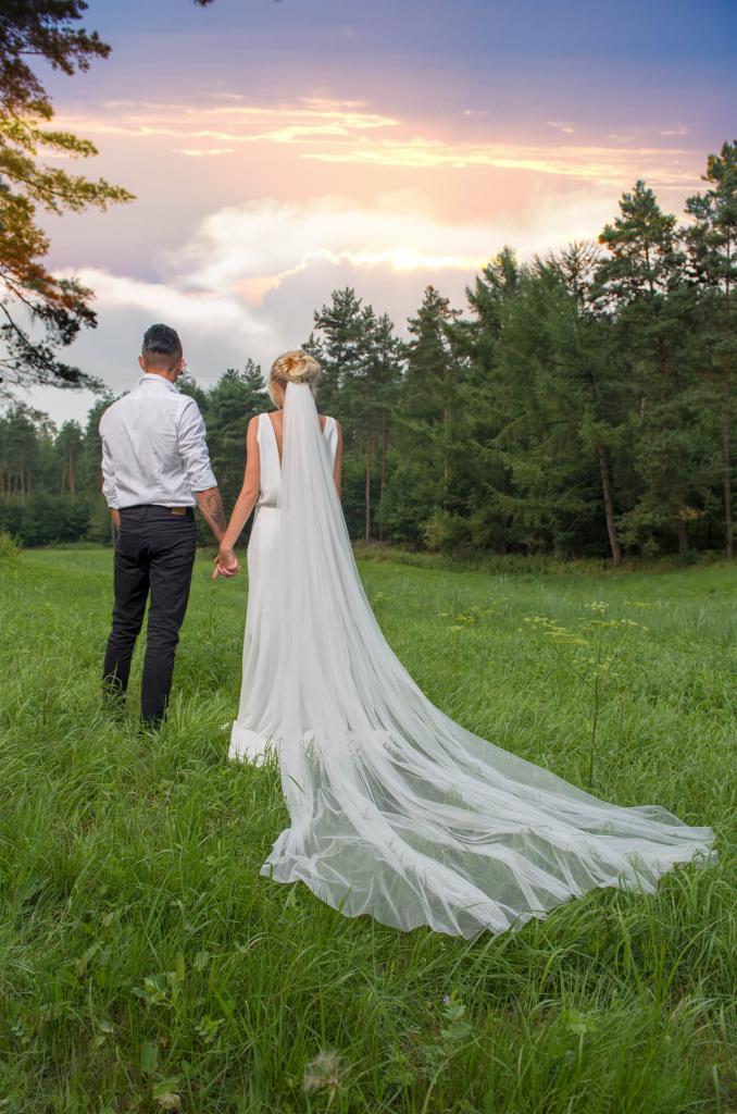 Para młoda, on w czarnych spodniach i białej koszuli, ona w białej sukni z długim welonem, trzymają się za ręce na łące, w tle las i zachodzące słońce.