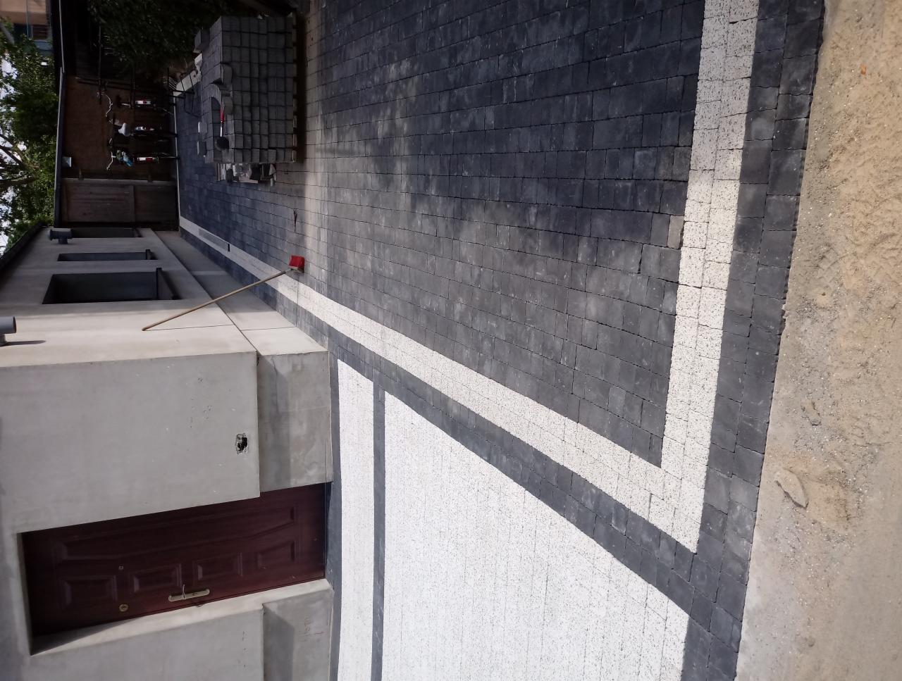 Ułożona szara kostka brukowa z białymi akcentami tworzącymi wzór, widoczne zapasy kostki i szczotka na kiju, surowy budynek w tle.