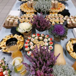 Stół cateringowy z różnorodnymi przekąskami, dekorowany wrzosami w doniczkach i dynią piżmową, z widocznymi koreczkami, roladkami i tartaletkami.