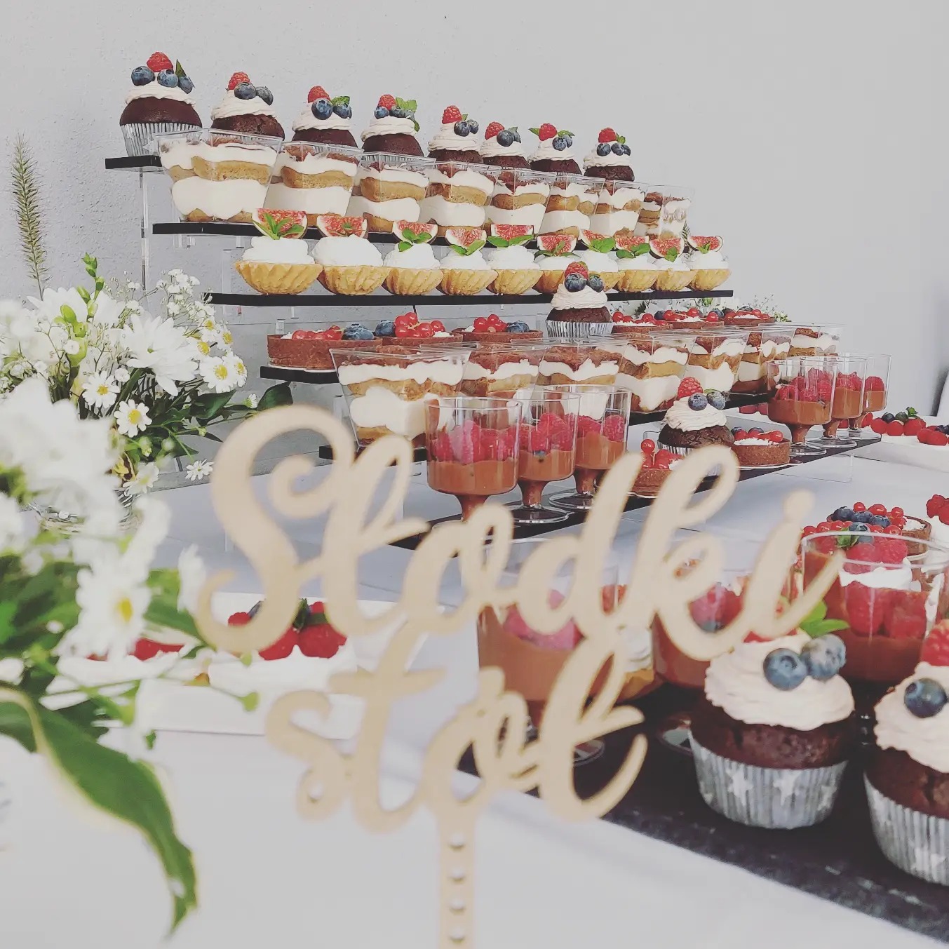 Bogato zastawiony stół z deserami: babeczki z owocami, desery w szklankach, tartaletki z figami, kompozycje kwiatowe, dekoracja 'Słodki stół'.