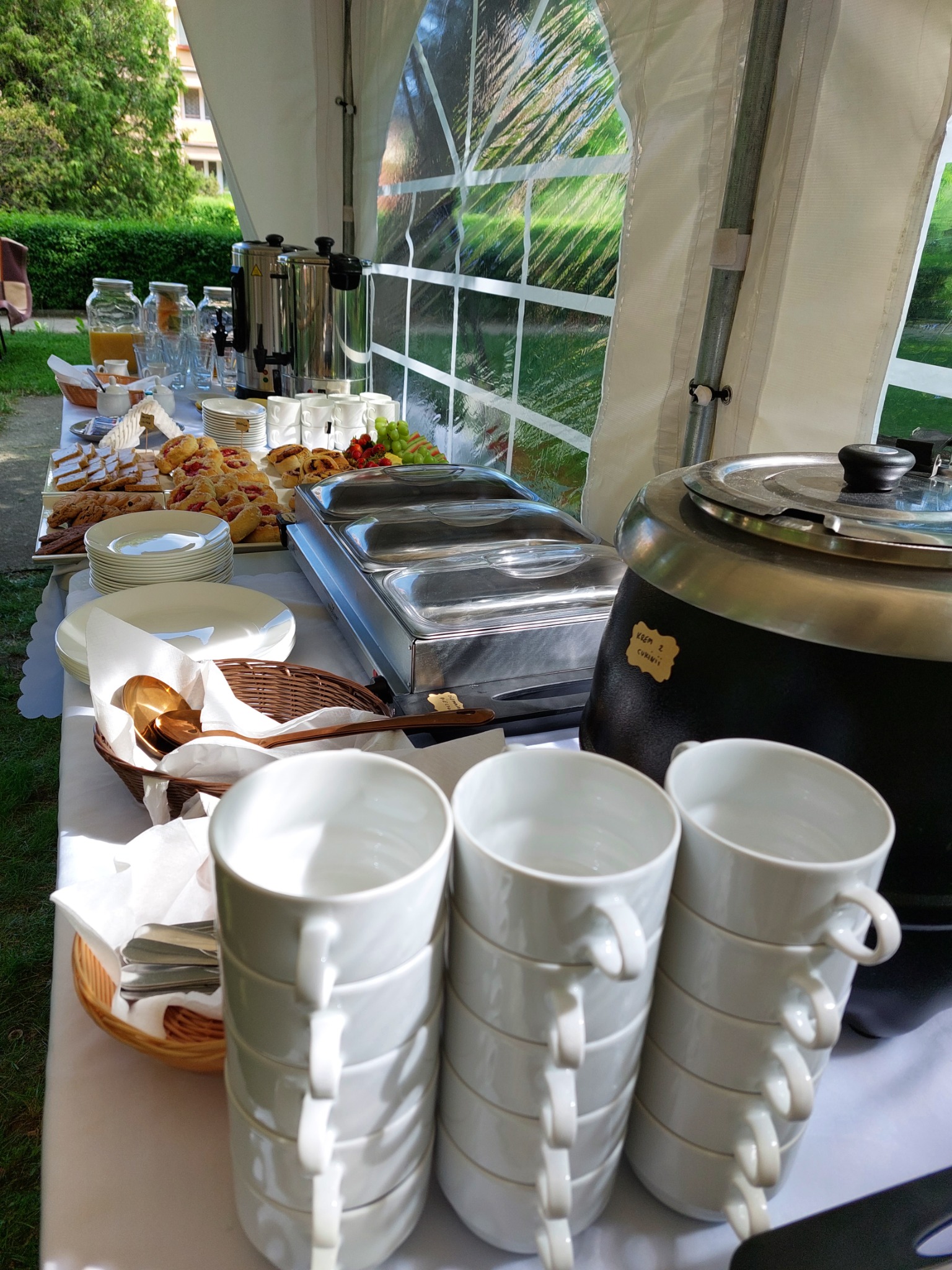 Stół cateringowy pod namiotem z widocznymi tacami z przekąskami, dzbankami z sokiem, oraz stosem białych filiżanek, zupa krem z cukinii w termosie.