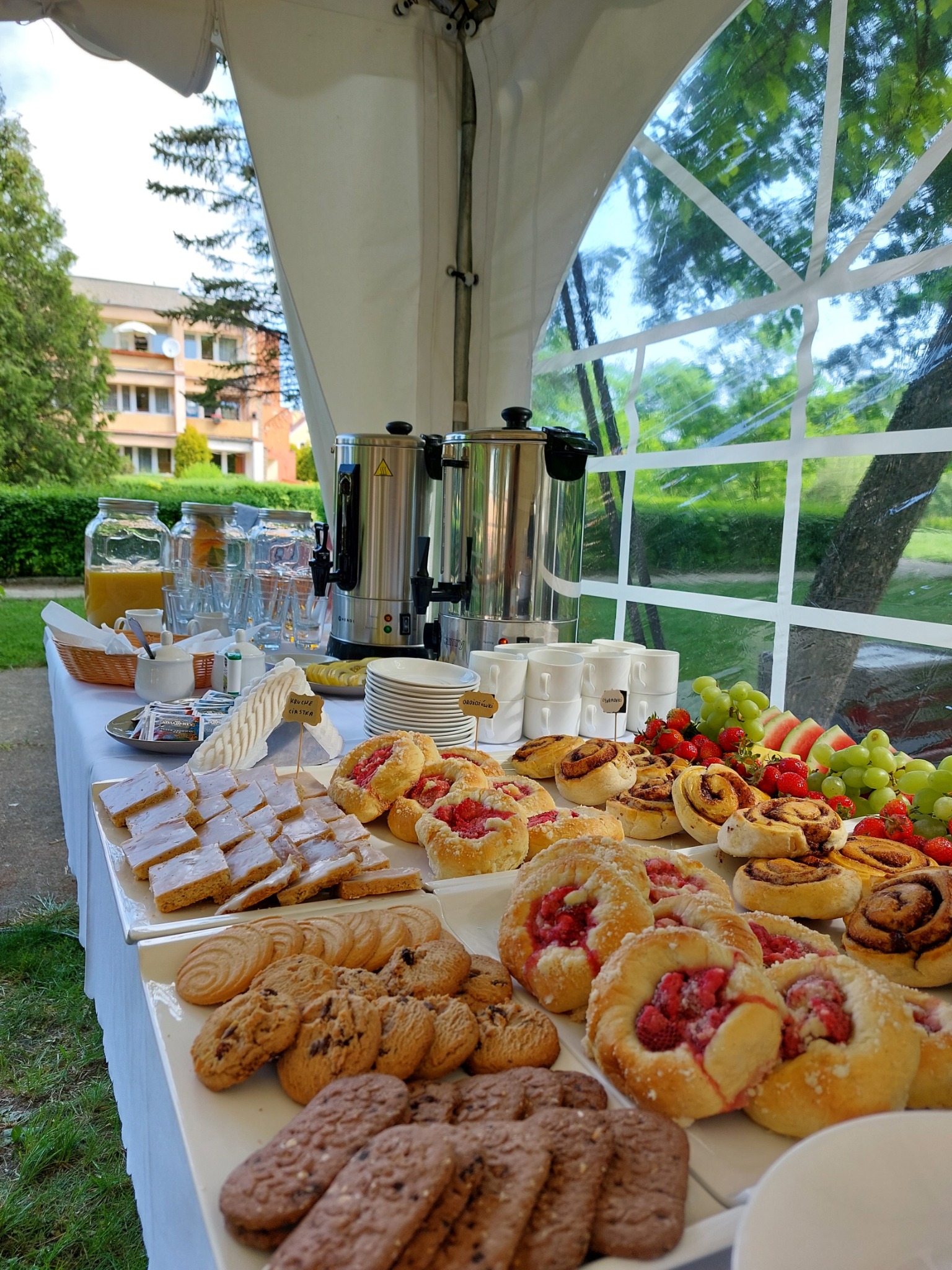 Stół cateringowy pod namiotem, pełen różnorodnych wypieków, owoców i napojów, z dwoma metalowymi dystrybutorami do napojów w tle, z widokiem na budynek i zieleń przez przezroczyste ściany namiotu.