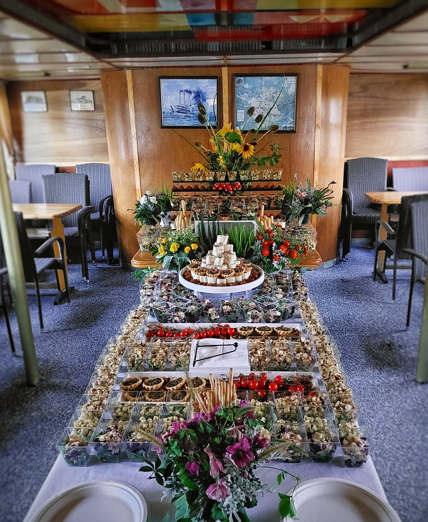 Bogato zastawiony stół cateringowy na statku, z piramidą przystawek w przezroczystych pojemnikach, dekoracjami kwiatowymi i grissini, na tle drewnianych paneli i marynistycznych obrazów.
