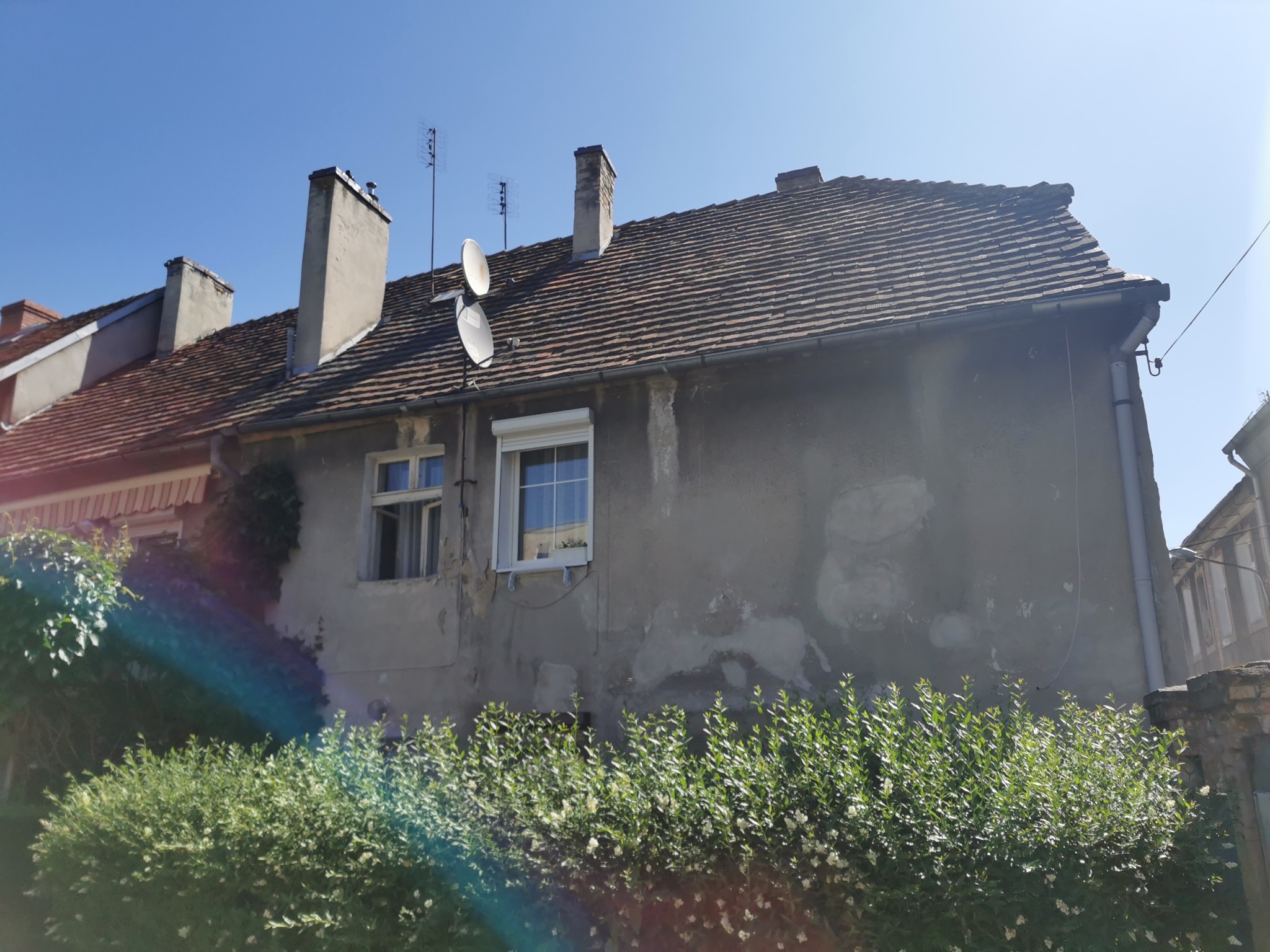 Dwukondygnacyjny budynek z szarego tynku z antenami satelitarnymi i oknami z roletami, otoczony zielenią. Widok z dołu, błękitne niebo w tle.