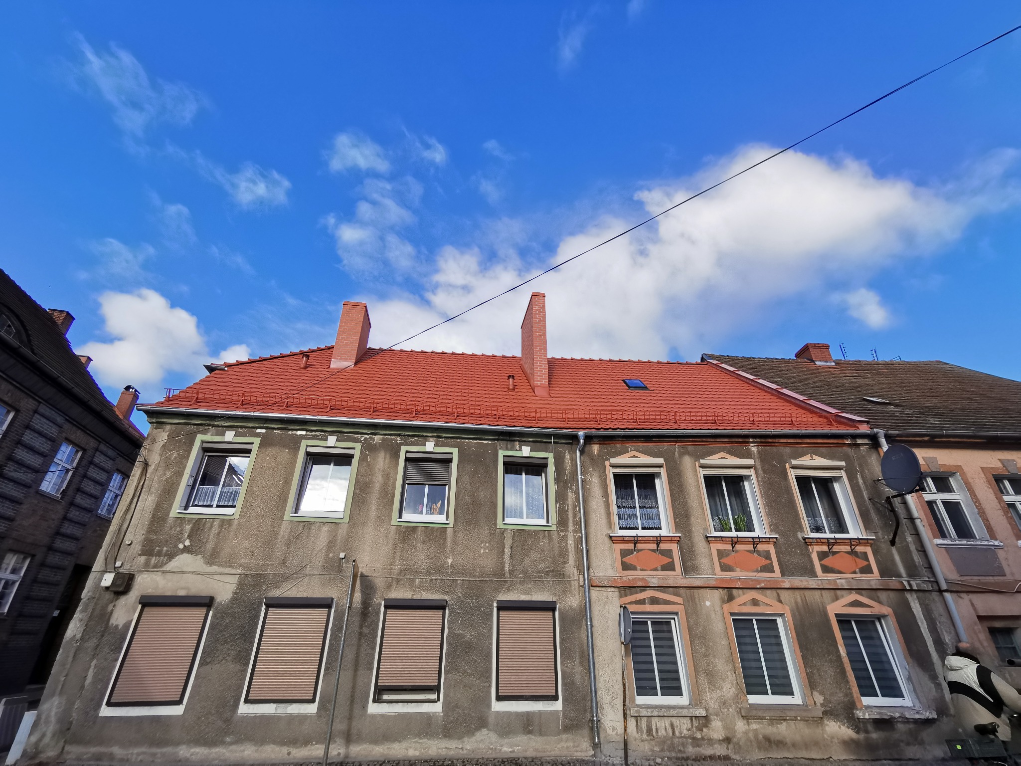 Fasada dwupiętrowego budynku z czerwonym dachem na tle błękitnego nieba z chmurami. Widoczne okna z roletami i ozdobnymi obramowaniami. Budynek o zróżnicowanej kolorystyce elewacji.