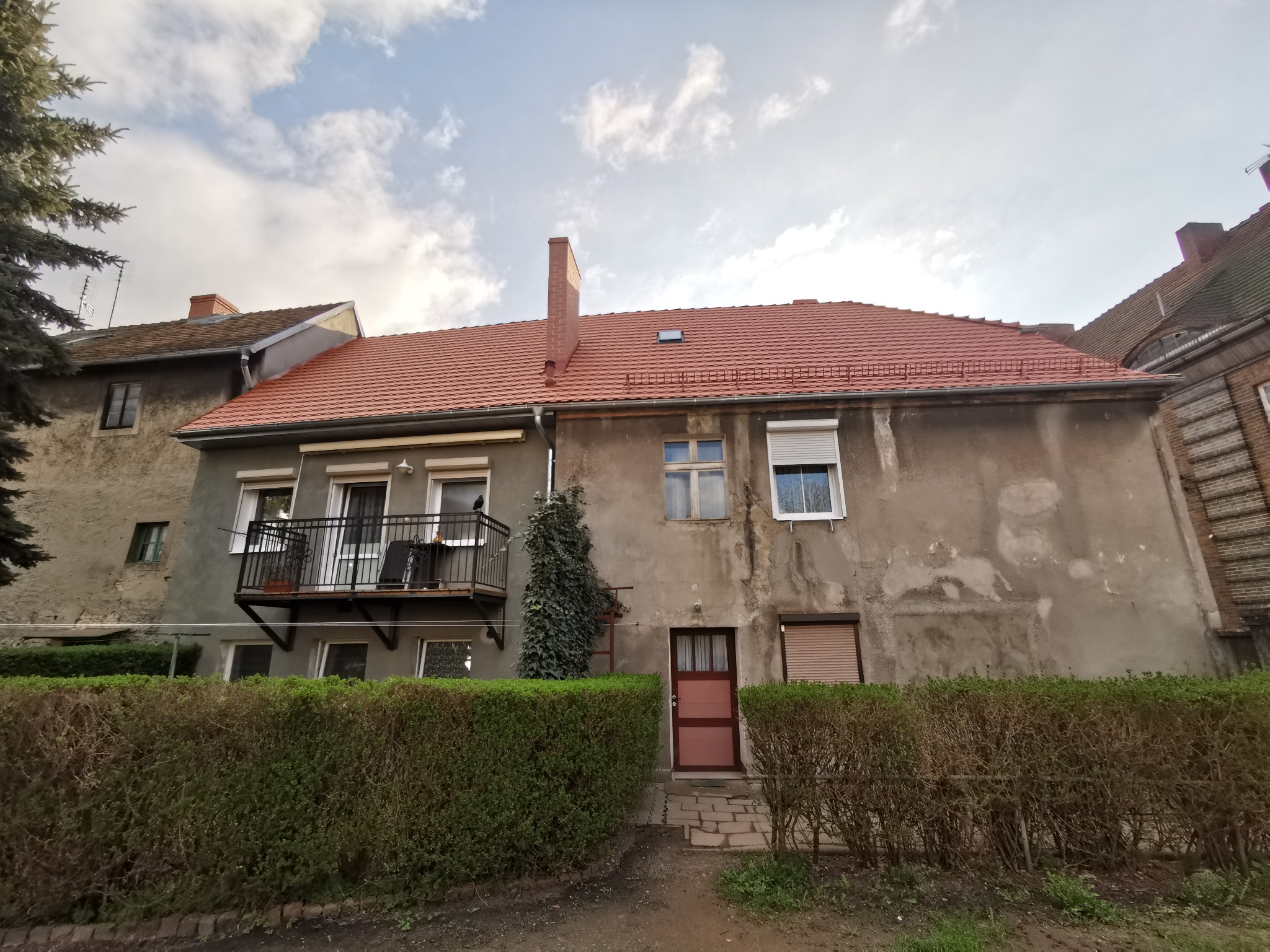 Dwupiętrowy dom z balkonem, czerwonym dachem i ceglanym kominem, otoczony zielonym żywopłotem. Widoczne ślady napraw na elewacji i częściowo opuszczone rolety okienne.