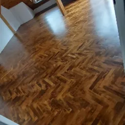 Błyszcząca, odnowiona podłoga parkietowa ułożona w jodełkę w jasnym poddaszu z oknami dachowymi i wyeksponowanymi belkami stropowymi.