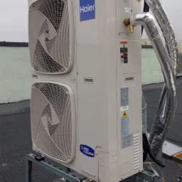 Zewnętrzna jednostka klimatyzacji Haier na dachu, zamontowana na metalowej konstrukcji, z widocznymi rurami w izolacji termicznej i elementami instalacji.