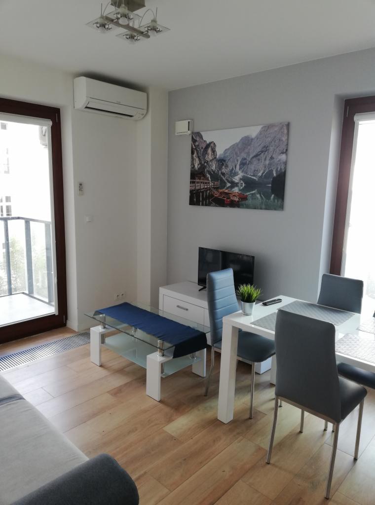 Wnętrze salonu z zamontowanym klimatyzatorem ściennym, widok na stół jadalny, telewizor i stolik kawowy z niebieskim bieżnikiem.