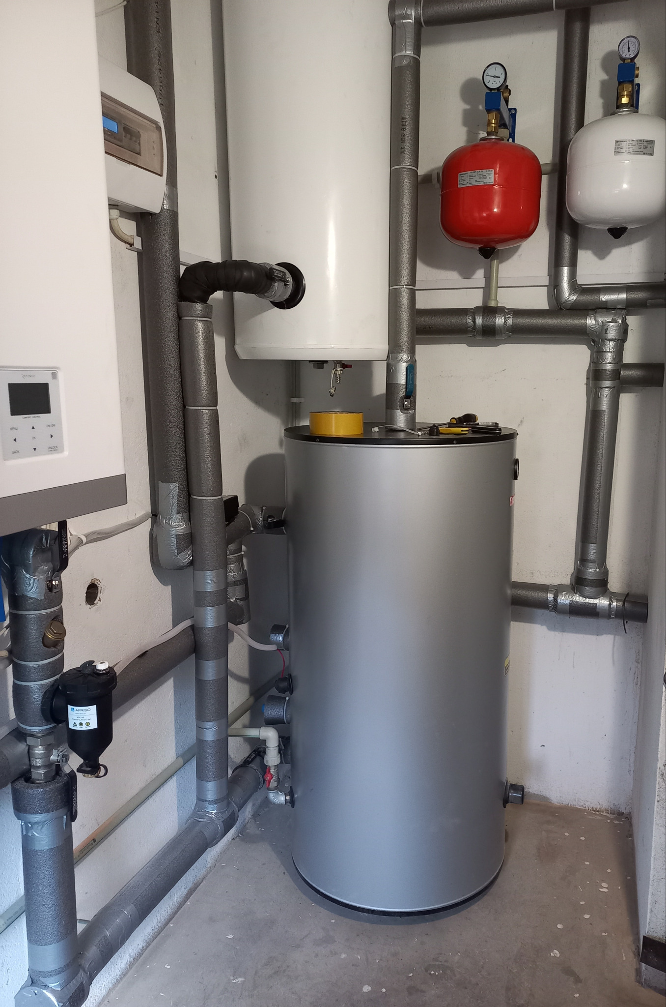 Szary, cylindryczny zasobnik ciepłej wody z żółtą taśmą na górze, otoczony rurami z izolacją, w pomieszczeniu technicznym z białymi ścianami i betonową podłogą.