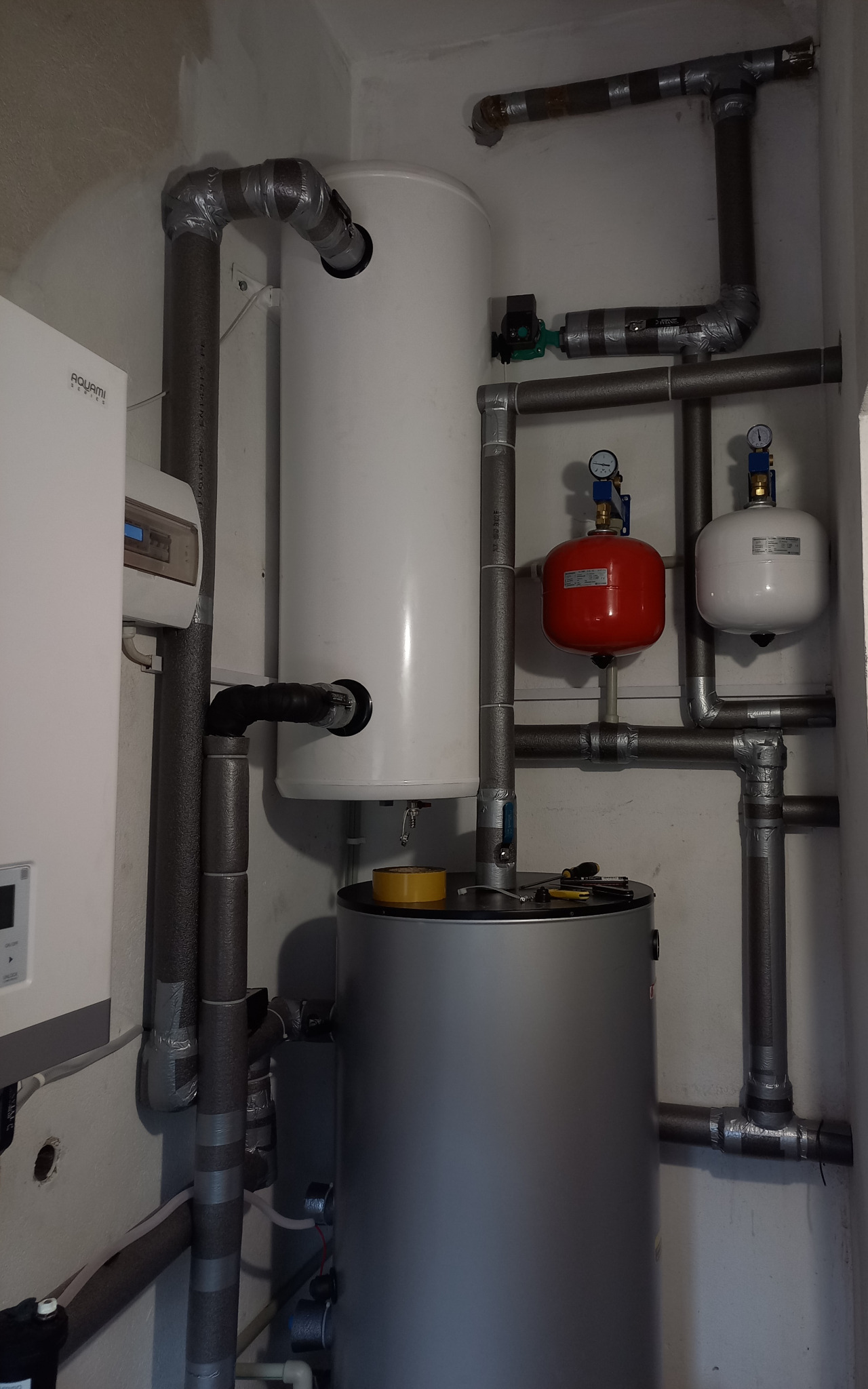 Instalacja pompy ciepła w kotłowni: widoczny biały zasobnik ciepłej wody, szary zbiornik buforowy, czerwone i białe naczynia przeponowe oraz rury z izolacją owiniętą szarą taśmą, na górze zbiornika...