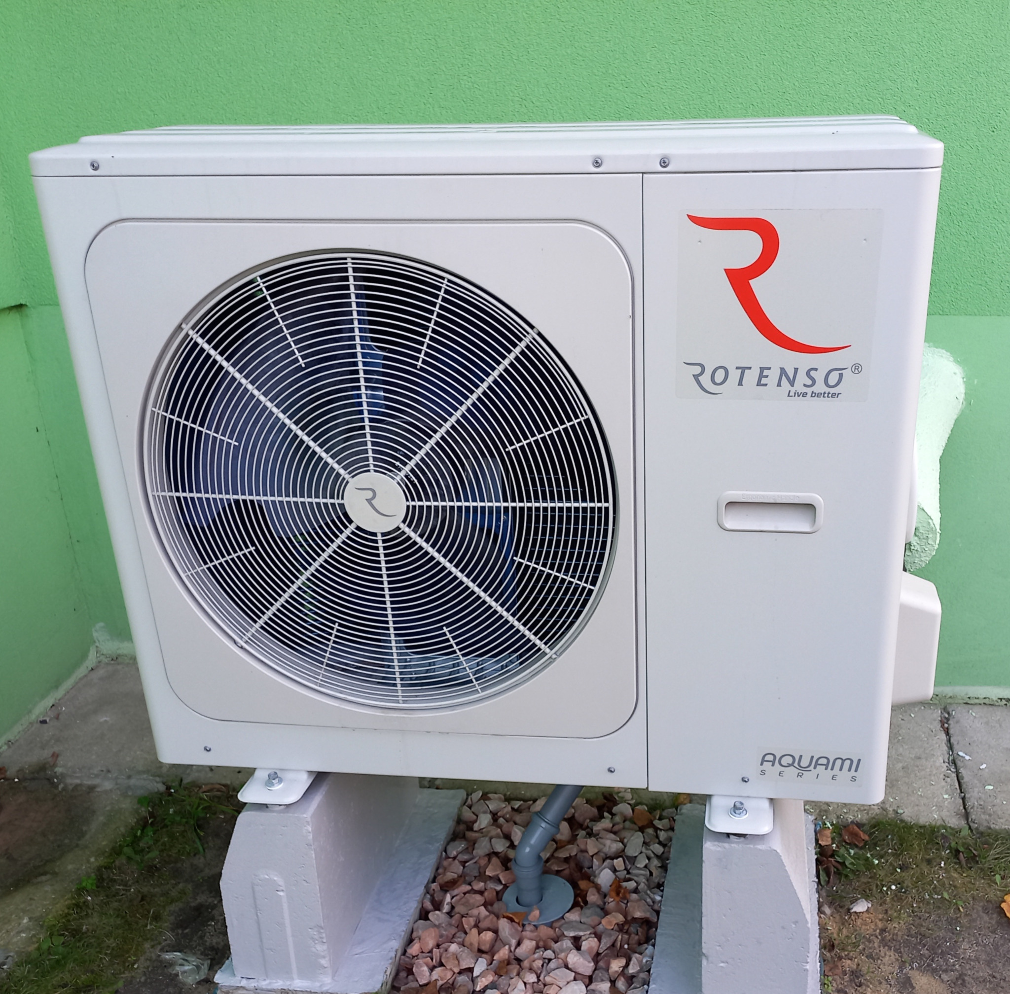 Zewnętrzna jednostka klimatyzacyjna Rotenso Aquami Series na betonowych podstawach, z widocznym odpływem skroplin, umieszczona przy zielonej ścianie budynku.