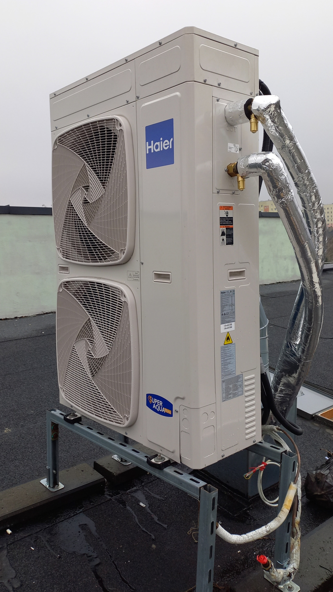 Zewnętrzna jednostka klimatyzacji Haier na dachu, zamontowana na metalowej konstrukcji, z widocznymi rurami w izolacji termicznej i elementami instalacji.