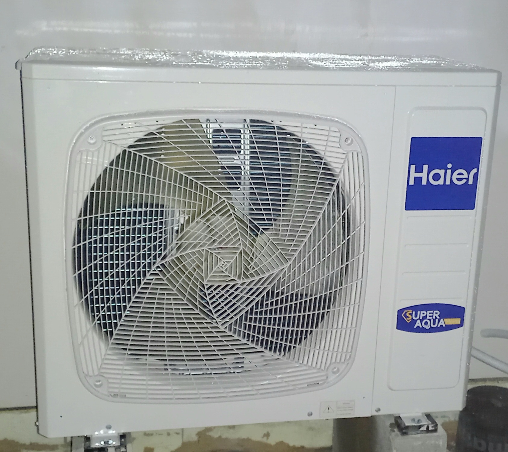 Zewnętrzna jednostka klimatyzacji Haier Super Aqua z folią ochronną na górze, zamontowana na betonowych wspornikach przy ścianie.