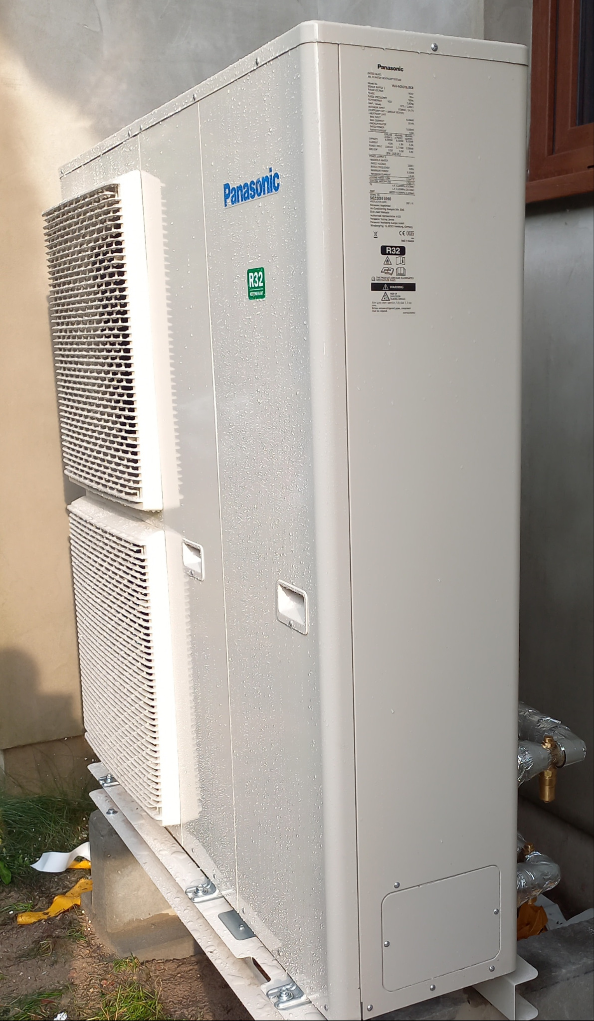 Zewnętrzna jednostka pompy ciepła Panasonic w kolorze jasnoszarym, z widocznymi lamelami wentylacyjnymi i naklejką R32, zamontowana na betonowej podstawie, lekko wilgotna od deszczu.