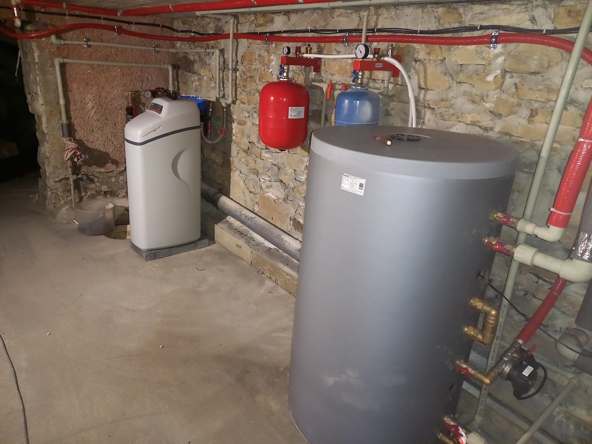 Instalacja w piwnicy z szarą butlą, czerwonym i niebieskim zbiornikiem wyrównawczym, oraz zmiękczaczem wody, widoczne rury miedziane i plastikowe.