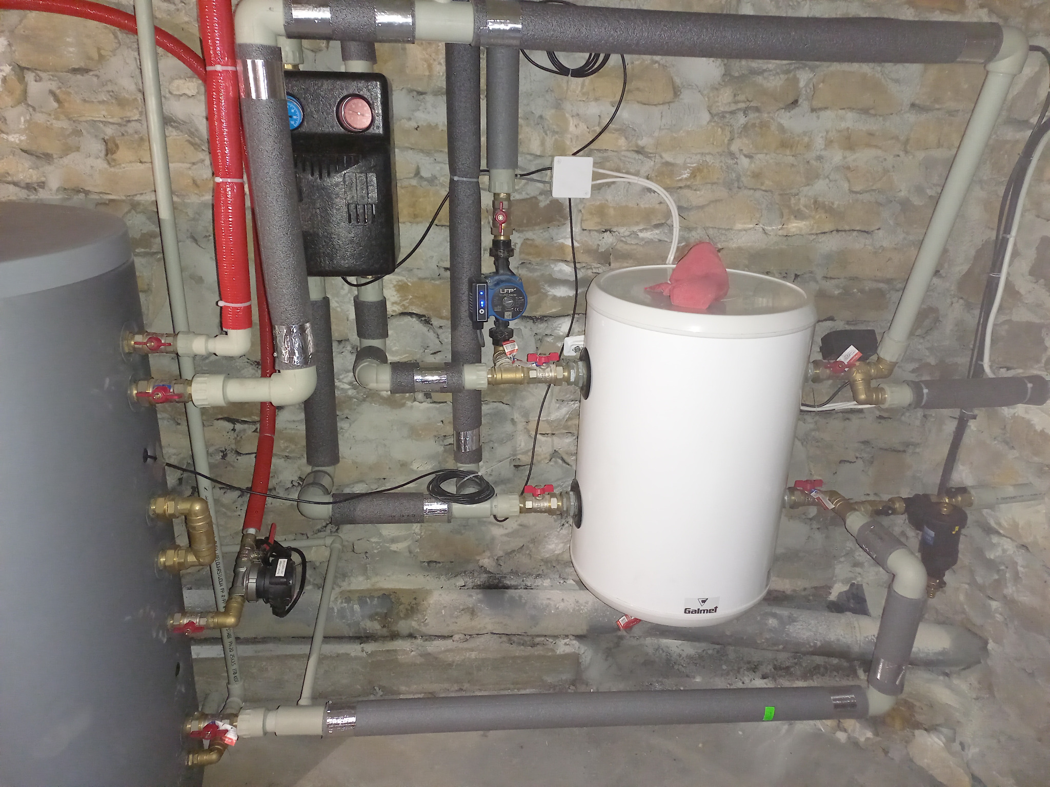 Instalacja dwóch zbiorników akumulacyjnych z rurami w izolacji termicznej, zaworami i pompą obiegową na tle ściany z kamienia.
