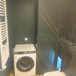 FRB Zbigniew Rzadzki - Nowoczesna łazienka z czarnymi płytkami imitującymi marmur, drewnopodobną podłogą, białą pralką Samsung i wiszącą toaletą.