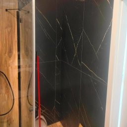 FRB Zbigniew Rzadzki - Kabina prysznicowa z czarnymi płytkami imitującymi marmur ze złotymi żyłami, drewnopodobne płytki na ścianie obok, czerwony mop w rogu.