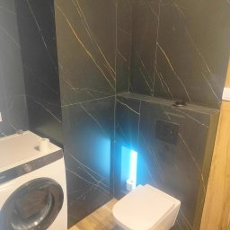 FRB Zbigniew Rzadzki - Nowoczesna łazienka z czarnymi płytkami imitującymi marmur, podwieszana toaleta z niebieskim podświetleniem, drewnopodobna podłoga i pralka w rogu.