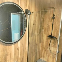 FRB Zbigniew Rzadzki - Nowoczesna kabina prysznicowa z czarną armaturą i okrągłym lustrem na ścianie wyłożonej drewnopodobnymi płytkami, widoczne niebieskie rurki instalacyjne.
