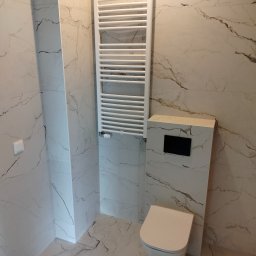 FRB Zbigniew Rzadzki - Nowoczesna łazienka wykończona białymi płytkami imitującymi marmur, z podtynkową toaletą, grzejnikiem drabinkowym i minimalistycznym włącznikiem światła.