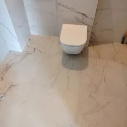 Wisząca toaleta z białą ceramiką, zamontowana na ścianie wyłożonej płytkami imitującymi marmur, z identyczną podłogą. Widoczne pionowe podświetlenie LED w narożniku ściany.