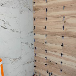 FRB Zbigniew Rzadzki - Montaż płytek imitujących drewno na ścianie z użyciem niebieskich krzyżyków dystansowych, widoczna także ściana z płytek imitujących marmur i fragment instalacji wodnej.