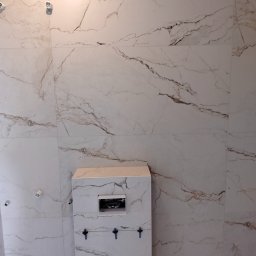 FRB Zbigniew Rzadzki - Ściana i zabudowa WC w łazience wyłożone białymi płytkami imitującymi marmur, widoczne niepodłączone punkty oświetleniowe i nieukończona instalacja