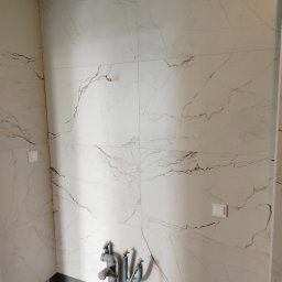 FRB Zbigniew Rzadzki - Ściana łazienki wyłożona białymi płytkami imitującymi marmur z widocznymi rurami instalacji wodnej i gniazdkami elektrycznymi.