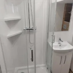 Nowoczesna, biała łazienka z kabiną prysznicową, umywalką z szafką i lustrem. Na ścianach białe płytki, w kabinie prysznicowej wbudowana półka narożna.