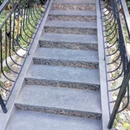 Schody zewnętrzne wyłożone szarymi płytkami z ciemnym wzorem, z czarną, ozdobną balustradą z elementami w kolorze złota, otoczone zielenią.