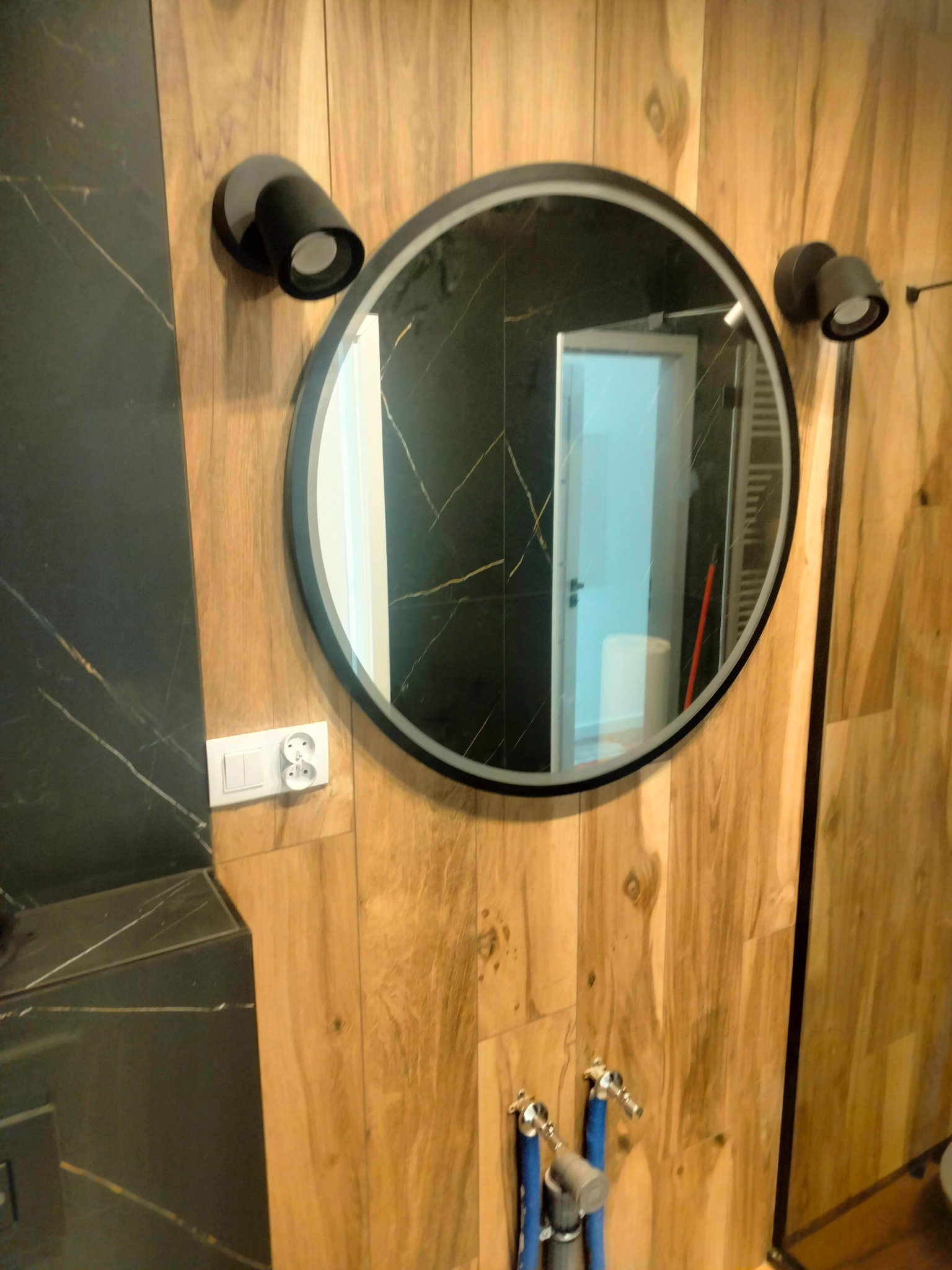 Okrągłe lustro z czarną ramą zawieszone na ścianie wyłożonej panelami imitującymi drewno, oświetlone dwoma czarnymi reflektorami; w odbiciu widać fragment łazienki z grzejnikiem drabinkowym.
