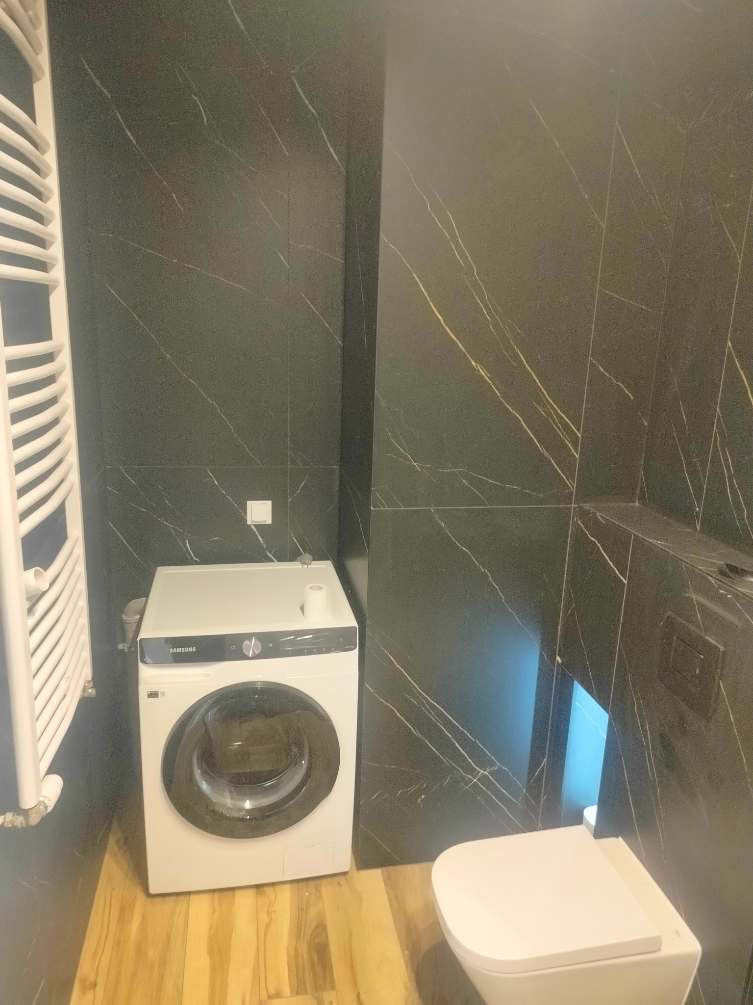 Nowoczesna łazienka z czarnymi płytkami imitującymi marmur, drewnopodobną podłogą, białą pralką Samsung i wiszącą toaletą.