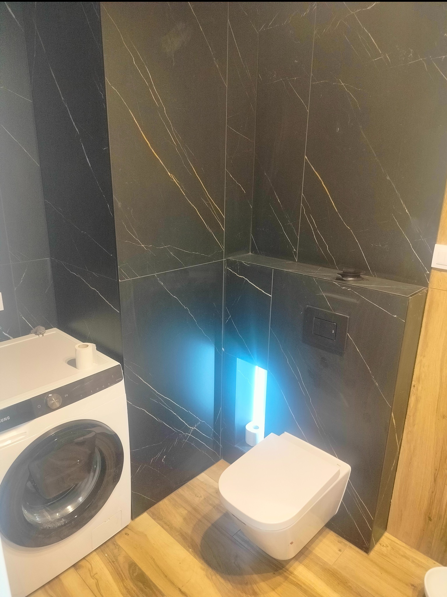 Nowoczesna łazienka z czarnymi płytkami imitującymi marmur, podwieszana toaleta z niebieskim podświetleniem, drewnopodobna podłoga i pralka w rogu.