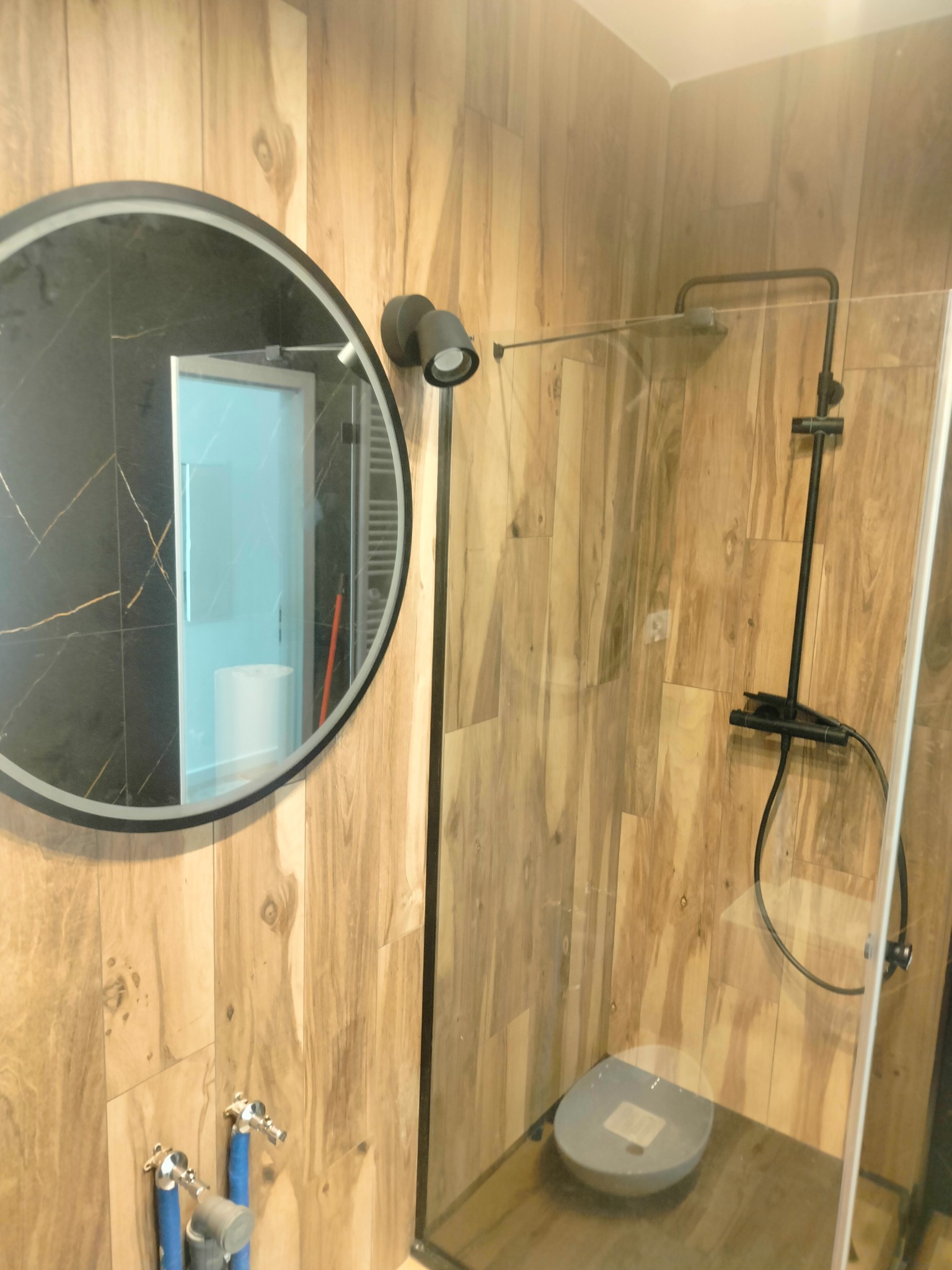 Nowoczesna kabina prysznicowa z czarną armaturą i okrągłym lustrem na ścianie wyłożonej drewnopodobnymi płytkami, widoczne niebieskie rurki instalacyjne.