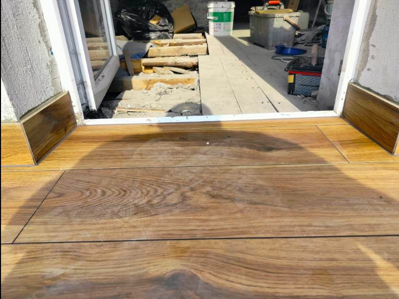 Ułożona podłoga z płytek imitujących drewno, widoczne łączenia, przy progu drzwi, w tle materiały budowlane i narzędzia.