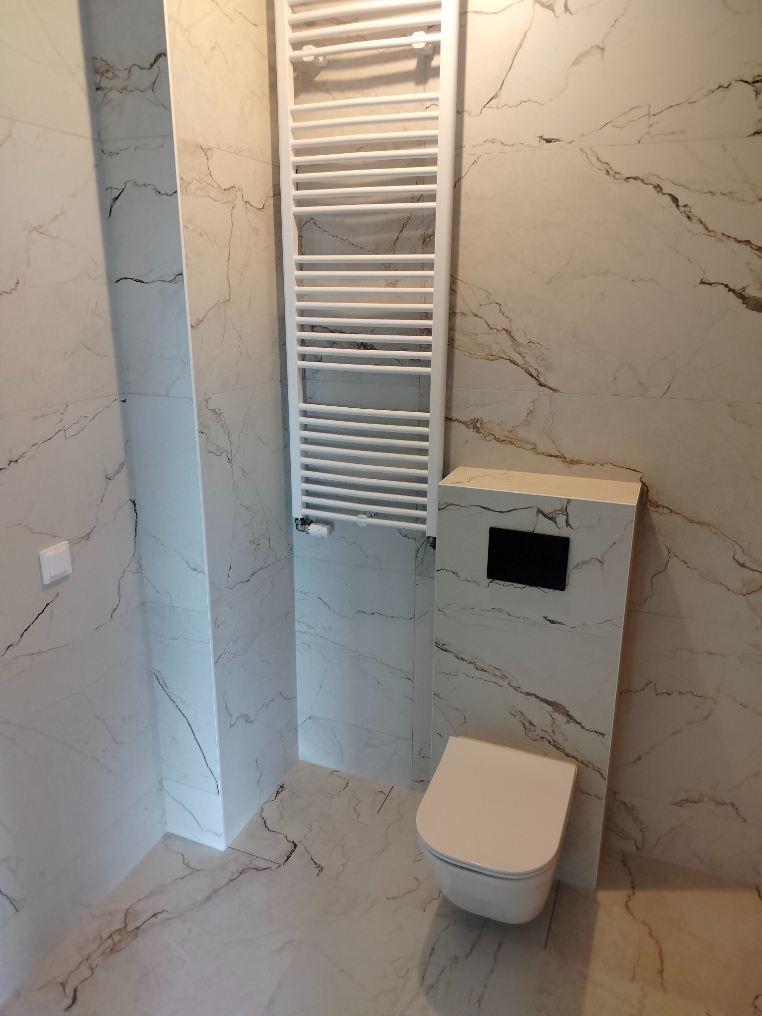 Nowoczesna łazienka wykończona białymi płytkami imitującymi marmur, z podtynkową toaletą, grzejnikiem drabinkowym i minimalistycznym włącznikiem światła.