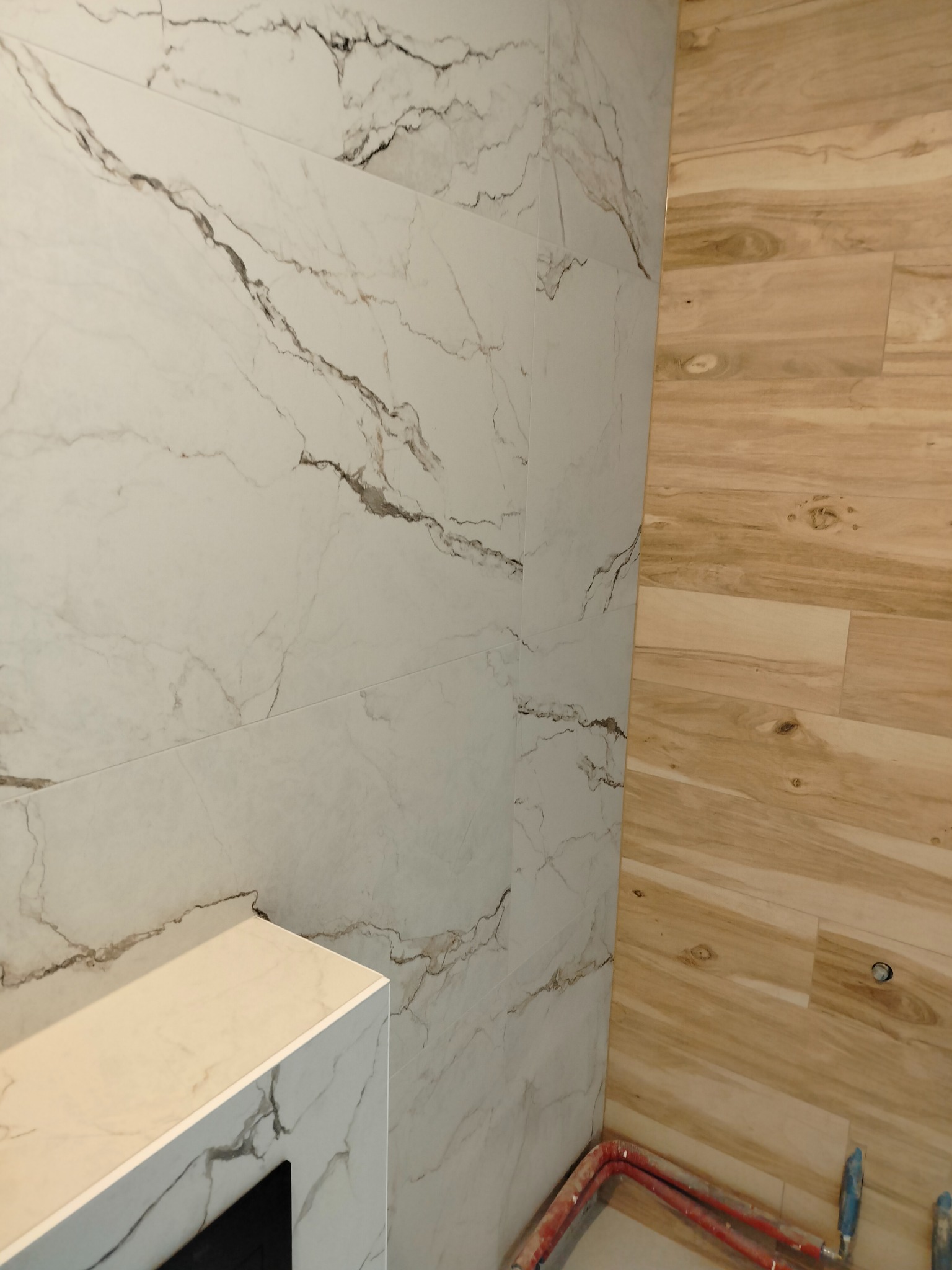 Ściana wyłożona płytkami imitującymi marmur z ciemnymi żyłami, sąsiadująca z panelami ściennymi imitującymi drewno. W dolnej części widoczne czerwone rury instalacji.