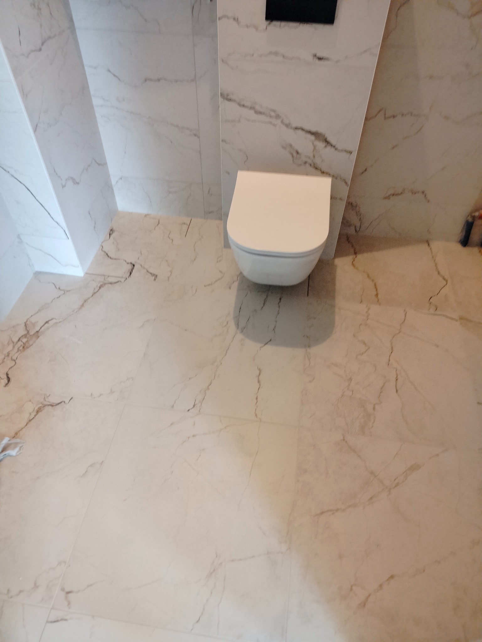 Wisząca toaleta z białą ceramiką, zamontowana na ścianie wyłożonej płytkami imitującymi marmur, z identyczną podłogą. Widoczne pionowe podświetlenie LED w narożniku ściany.