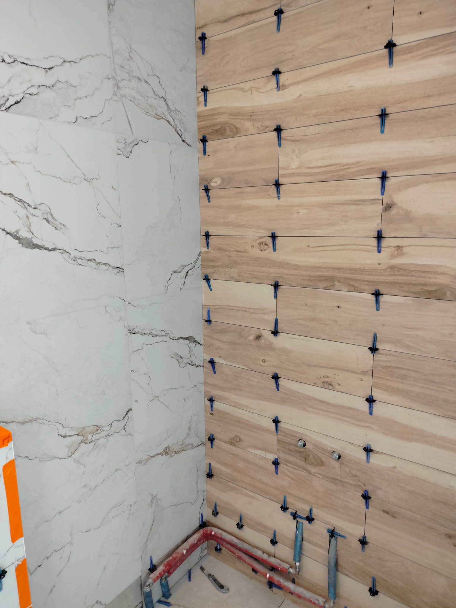 Montaż płytek imitujących drewno na ścianie z użyciem niebieskich krzyżyków dystansowych, widoczna także ściana z płytek imitujących marmur i fragment instalacji wodnej.