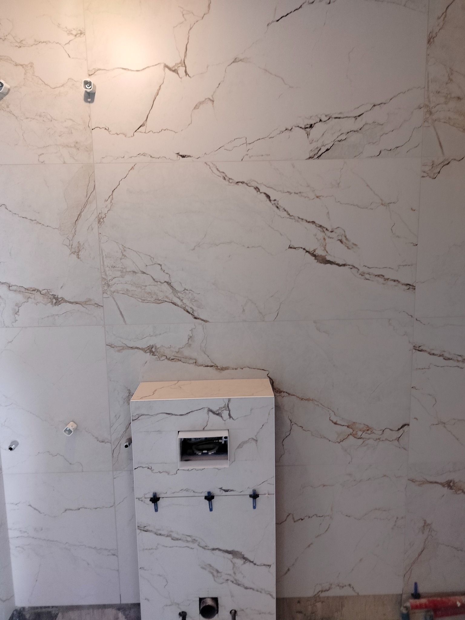 Ściana i zabudowa WC w łazience wyłożone białymi płytkami imitującymi marmur, widoczne niepodłączone punkty oświetleniowe i nieukończona instalacja