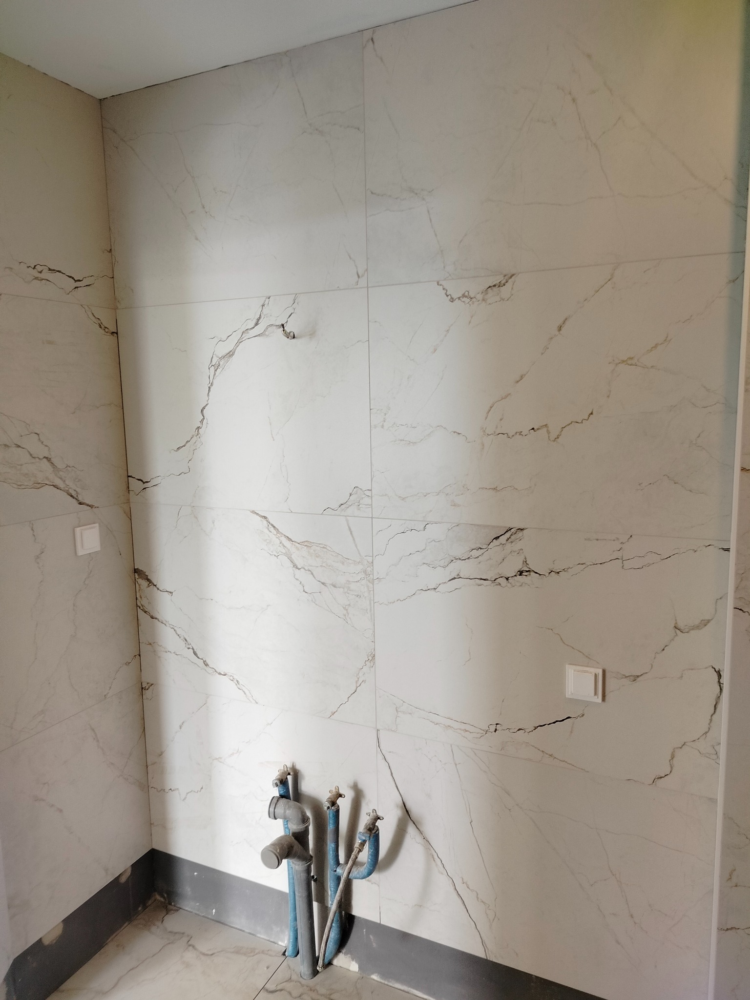 Ściana łazienki wyłożona białymi płytkami imitującymi marmur z widocznymi rurami instalacji wodnej i gniazdkami elektrycznymi.