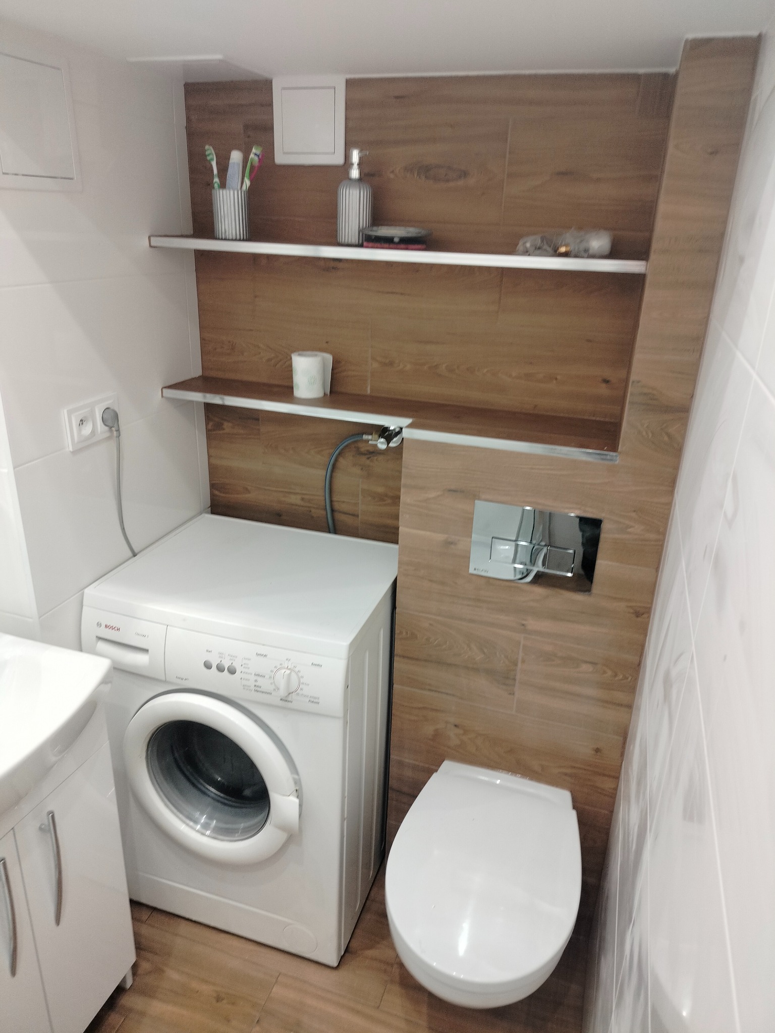 Mała łazienka z białymi i drewnopodobnymi płytkami, pralką Bosch, podwieszaną toaletą i półkami z akcesoriami.
