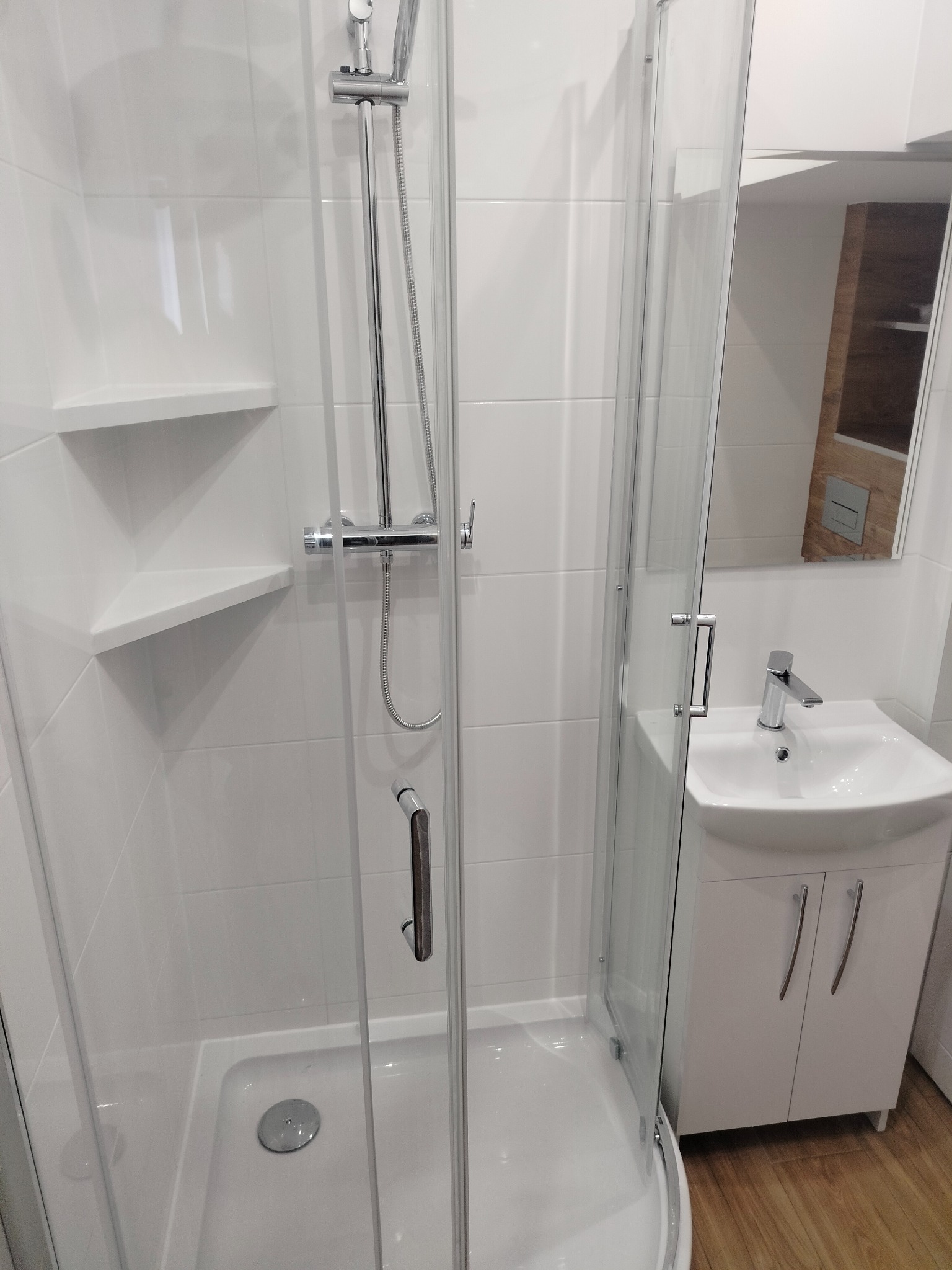 Nowoczesna, biała łazienka z kabiną prysznicową, umywalką z szafką i lustrem. Na ścianach białe płytki, w kabinie prysznicowej wbudowana półka narożna.