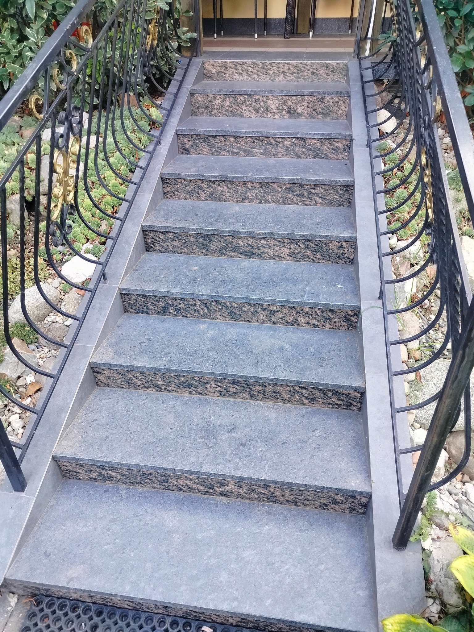 Schody zewnętrzne wyłożone szarymi płytkami z ciemnym wzorem, z czarną, ozdobną balustradą z elementami w kolorze złota, otoczone zielenią.