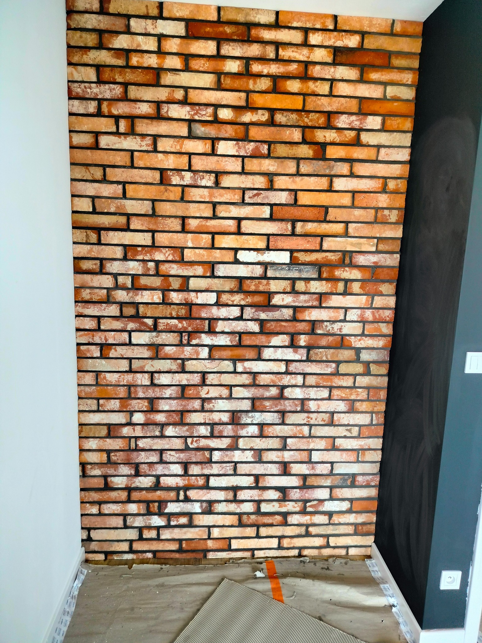 Ściana wyłożona cegłą klinkierową w odcieniach pomarańczy i beżu, z ciemną fugą, sąsiadująca z białą i grafitową ścianą. Na podłodze widoczne płyty ochronne i pomarańczowa taśma malarska.