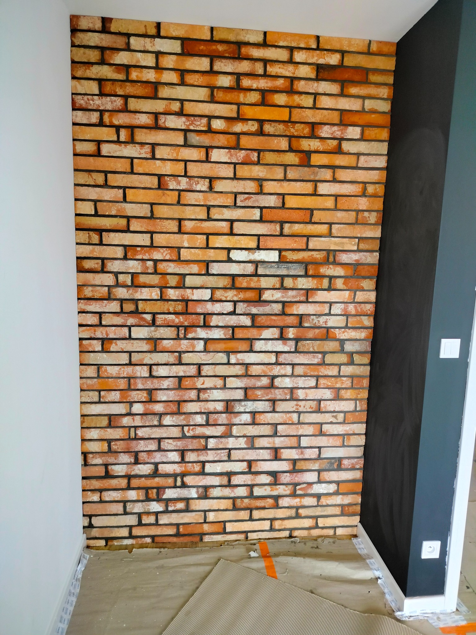 Ściana z cegły klinkierowej w odcieniach pomarańczy i bieli, fragment białej ściany oraz ciemnoszara ściana z elementami wykończenia, zabezpieczona podłoga kartonem falistym.