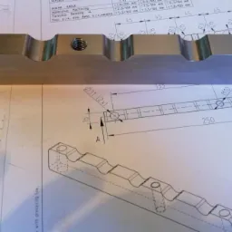 Precyzyjnie obrobiony metalowy element z gwintowanymi otworami, leżący na technicznym rysunku z wymiarami i przekrojami.