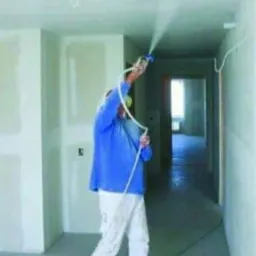 Pracownik w niebieskiej koszuli natryskuje białą substancję na sufit w pomieszczeniu z surowymi ścianami, używając pistoletu natryskowego podłączonego do węża.